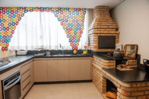 Área gourmet da Casa Caiçara com churrasqueira de alvenaria e fogão a lenha em tijolos aparentes, bancada ampla e coifa metálica, executada pela Mazí Construtora.